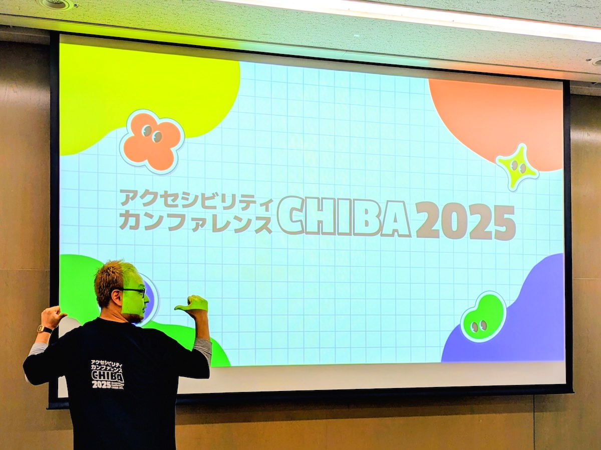 スクリーンに映し出されたアクセシビリティカンファレンス千葉2025のロゴとスタッフTシャツの背面のロゴを見せているmasuP9