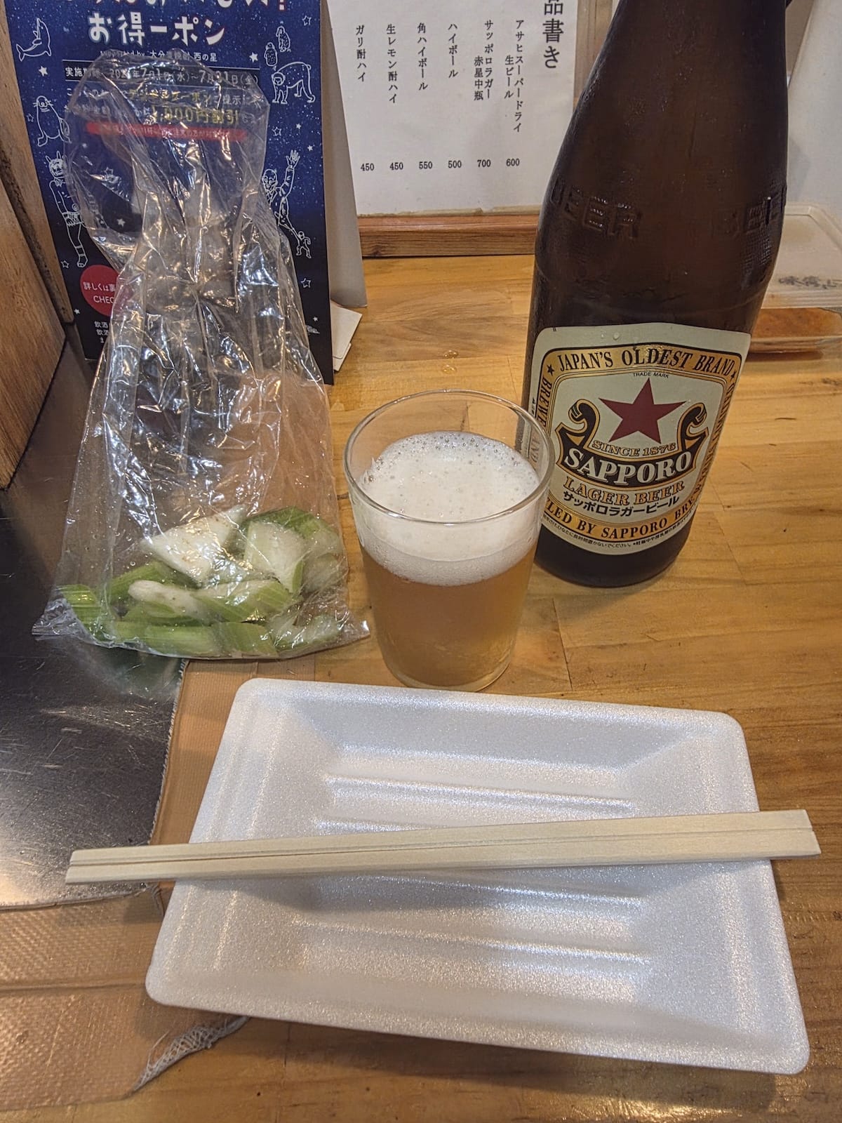 立ち飲みのカウンター。サッポロラガーの中瓶とビールの入ったグラス、ビニール袋に入ったセロリの浅漬け、割り箸を載せた発泡スチロールのトレイが並び、奥の壁には手書きの品書きが貼ってある