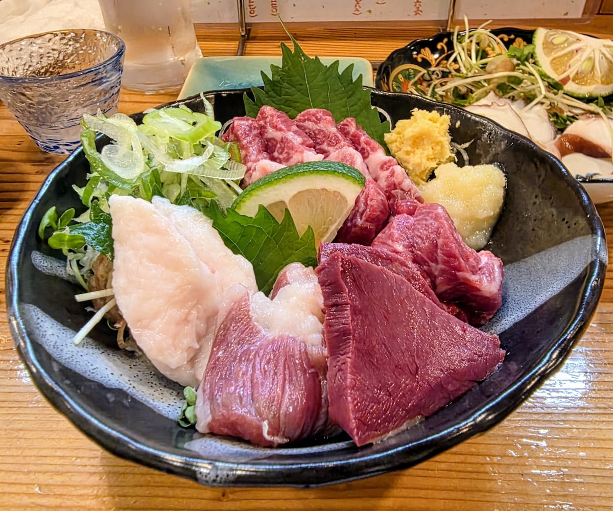 黒い大皿に盛られた馬刺しの盛り合わせ。赤身、白い脂身、レバーのような濃い赤の部位が大葉の上に並び、おろし生姜とおろしにんにく、刻みネギ、すだちが添えられている