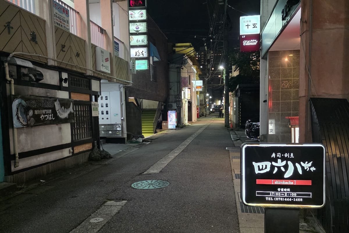 夜の歓楽街の細い路地。両側の建物に居酒屋やスナックの看板が並び、手前には寿司・割烹の「四六八ちゃ」という店の看板が立っている